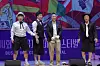 Four performers stand on a brightly lit stage in front of a colorful festival backdrop.