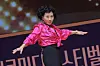 Performer in a bright pink blouse extends her arms on a stage with Korean lettering behind her.
