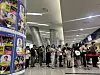 Crowd waiting in line inside a brightly lit terminal beside a poster-covered column.