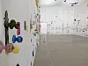 Colorful wall-mounted artworks line a white contemporary gallery beside an open exhibition space.
