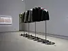 Dark garments hang from mobile metal stands in a minimalist art gallery.