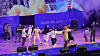 Ensemble dancers perform on a brightly lit purple stage with event signage in the background.