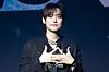 A male performer in black stands on a dark stage, smiling and forming a gesture with his hands.