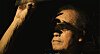 Close-up portrait of a person in dramatic shadow with one hand raised.