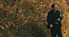 Man holding a smartphone while walking through a wooded area with autumn foliage.