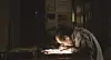 Person reading at a desk under a small pool of light in a dark room with shelves.