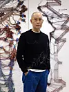 Man in black sweater standing before intricate wire and thread wall sculptures.