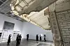 Visitors view a large suspended architectural installation in a bright museum gallery.