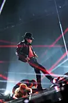 Hat-wearing musician performs near the stage edge amid red and blue concert lights.