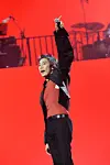 Performer singing with one arm raised on a concert stage with a red backdrop.