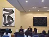 Two speakers sit before an audience in a modern room with abstract artwork on the walls.