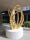 Abstract gold-bead loop sculpture displayed on a white pedestal in a modern courtyard.