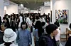 A busy art fair with visitors browsing gallery booths and a colorful painting displayed on the right.