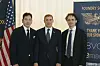 Three men in suits pose beside a U.S. flag and event sponsor backdrop.