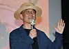 Lee Joon-ik, wearing a hat and glasses, speaks into a microphone with one hand raised.