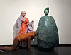 Costumed performer beside a large ornate green sculpture in a white gallery.