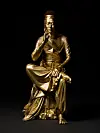 Gilded seated sculpture with ornate robes illuminated against a black background.