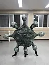 Abstract dark metal sculpture with rounded forms and pointed legs in a room with barred windows.