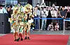 Costumed participants wearing elaborate gold crown outfits walk along a red carpet before a museum audience.