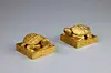 Two ornate gold-colored objects on square bases against a light gray background.