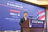 Man speaking at a podium before a Korean-American business forum backdrop with flags and event signage.