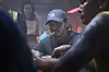 Man in a baseball cap seated indoors, partially surrounded by people in low light.