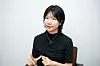 Seated person in a black top gestures during an interview against a light background.
