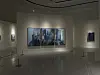Contemporary artworks hang in a softly lit gallery with a reflective floor and viewing barriers.