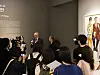 A speaker talks to a crowd in a gallery beside a colorful artwork.