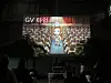 A large projection screen shows a presenter and audience above silhouetted attendees in a dark venue.