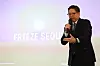 Patrick Lee speaks into a microphone before a projected Frieze Seoul backdrop.
