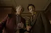 Two men in formal suits look down toward the camera in an ornate, dimly lit room.