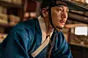 Ji Chang-wook as Cho Won wears blue traditional clothing and black headgear in a warmly lit wooden room.