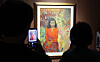 Silhouetted visitors view and photograph a brightly colored framed portrait in a dim exhibition gallery.