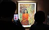 Silhouetted visitors view and photograph a brightly colored framed portrait in a dim exhibition gallery.