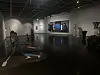 Dim gallery interior with a visitor and contemporary paintings and sculptures on display.