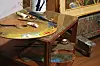 Artist’s palette, brushes and paint tubes on a wooden table beside framed artwork.