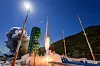 Rocket launching beside a service tower, with bright exhaust and mountains in the background.