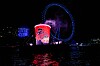 Red illuminated cup display beside a blue Ferris wheel at night, reflected on dark waterfront water.