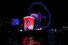 Red illuminated cup display beside a blue Ferris wheel at night, reflected on dark waterfront water.