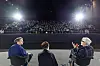 Three seated panelists address a large audience in a dimly lit auditorium.