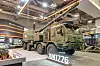 Green armored military vehicle on display inside a brightly lit exhibition hall.