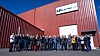 Large group posing outside a red industrial building with a company sign.