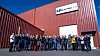 Large group posing outside a red industrial building with a company sign.