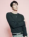 Young man in a dark sweater and jeans posing with folded arms against a pink background.