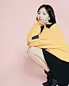 Seated woman in a yellow wrap and black boots against a pink studio background.