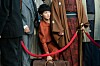 Woman in vintage-style clothing stands behind a red velvet rope among hanging garments.