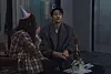Kim Young-kwang sits beside a small cake as a woman in a party hat faces him indoors.