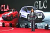 Presenter beside a silver Mercedes-Benz GLC SUV, with a red CLA on a lit exhibition stage.