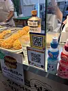Two QR payment signs beside prepared food trays and bottled drinks at a Seoul market stall.
